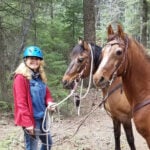 A person wearing a blue helmet and red jacket is standing in a forest holding the reins of two brown horses.