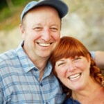 A man in a blue cap and plaid shirt smiles with a woman with red hair, both embracing and smiling outdoors.